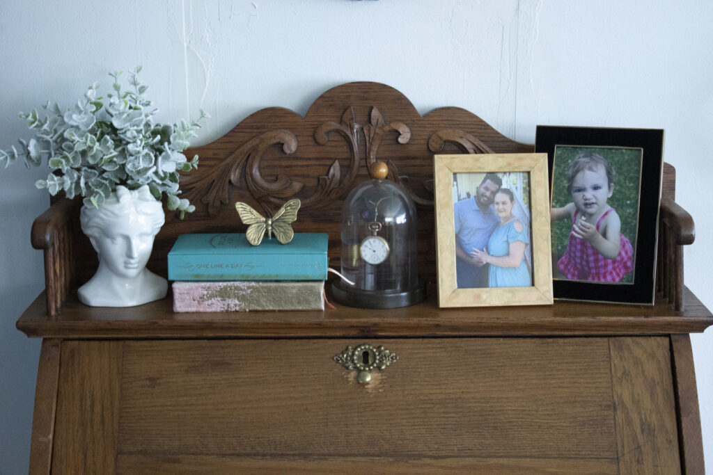 Vintage secretary desk styled with a bust, books, butterfly, two photo frames, and a vintage pocketwatch